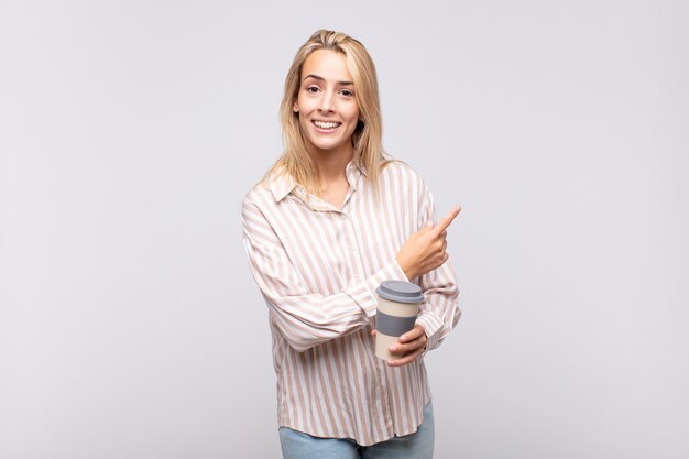 Mujer joven con un café mirando emocionado y sorprendido apuntando hacia un lado y hacia arriba para copiar el espacio
