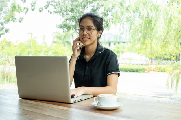 Mujer joven en cafetería usando una computadora portátil y hablando en un teléfono celular.