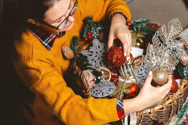 Mujer joven con una caja eligiendo adornos navideños en una canasta