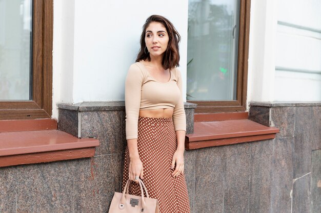 Foto mujer joven en la calle de la ciudad. una hermosa morena con estilo está de pie contra una pared blanca de la casa.