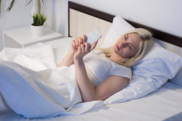 Mujer joven en la cama a altas horas de la noche enviando mensajes de texto con teléfono móvil cansado de quedarse dormido.