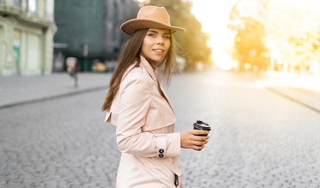 Mujer joven camina por las calles de la ciudad vestida con un sombrero y un impermeable. Café para llevar. copia espacio