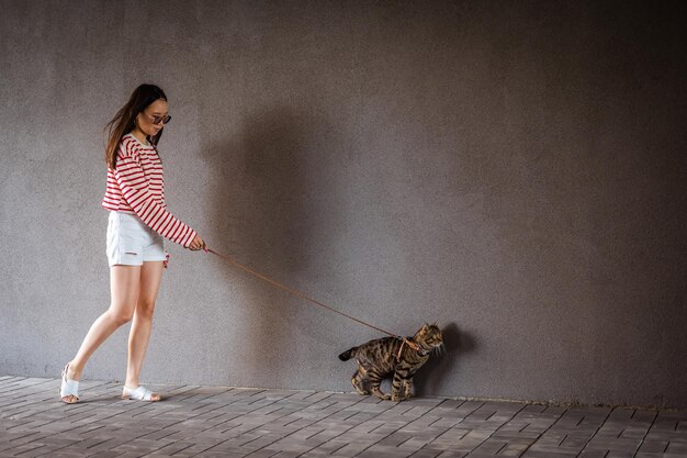 Una mujer joven camina con un gato tabby gris en una correa contra una pared gris