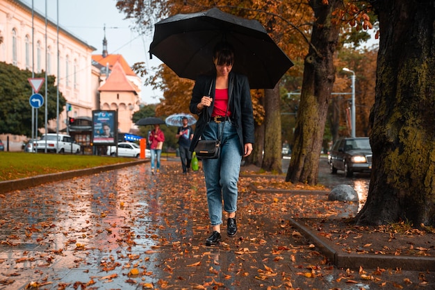 Mujer joven camina con paraguas negro bajo la lluvia. hojas amarillas en el suelo. el otoño está próximo