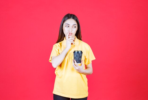 Mujer joven en camisa amarilla sosteniendo una taza de café desechable negra, pensando y teniendo una buena idea