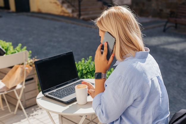 Mujer joven con una camisa azul está trabajando en una computadora portátil en un café y hablando por teléfono