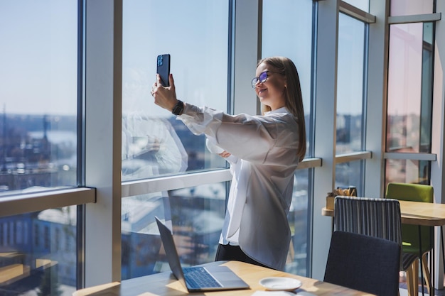 Una mujer joven con una camisa blanca y anteojos está de pie junto a la ventana con un teléfono y hablando por un enlace de video El gerente celebra una conferencia en línea