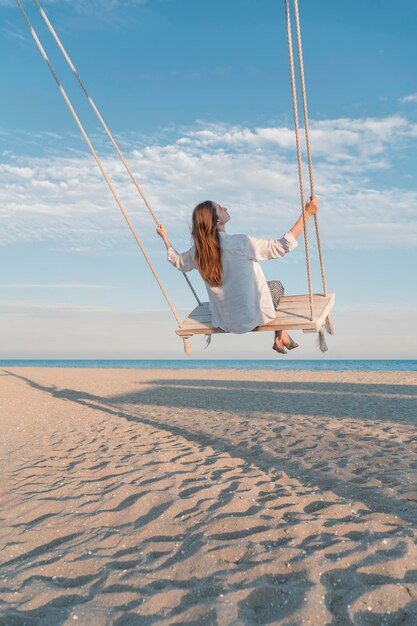 Mujer joven con camisa blanca en una cuerda balanceándose en el cielo azul y el fondo del mar chica feliz divirtiéndose balanceándose alto en el aire