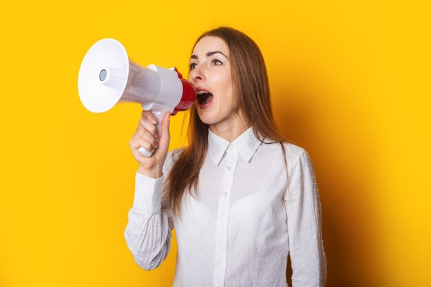 Mujer joven con una camisa blanca hace un anuncio en un megáfono sobre un fondo amarillo