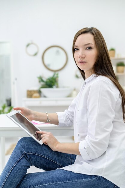 Una mujer joven con una camisa blanca y jeans se sienta en una mesa con una tableta en las manos Una gerente de oficina realiza un trabajo en un dispositivo moderno