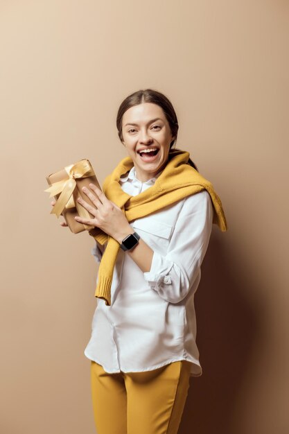 Mujer joven en camisa blanca con regalo de navidad hecho a mano empacado en papel kraft sobre fondo de pared epmty...