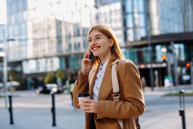 Mujer joven en camisa blanca con taza de café para llevar contestando una llamada telefónica mientras cruza cerca