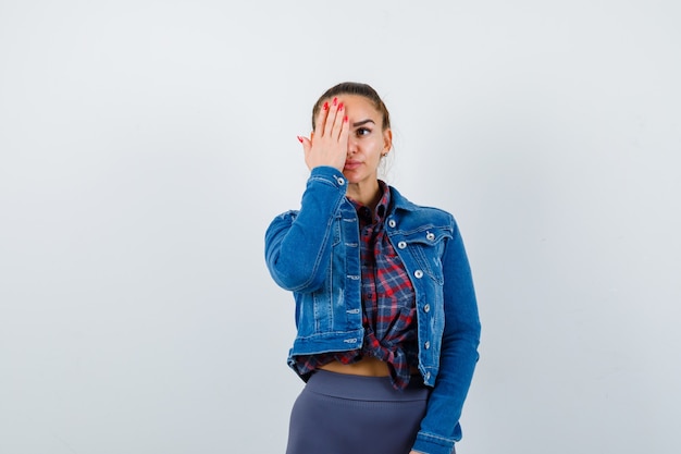 Mujer joven en camisa a cuadros, chaqueta, pantalón con la mano en el ojo y mirando serio, vista frontal.