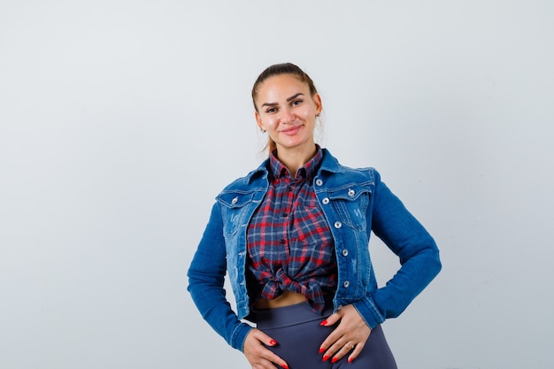 Mujer joven en camisa a cuadros, chaqueta, pantalón posando mientras está de pie y mirando encantador, vista frontal.