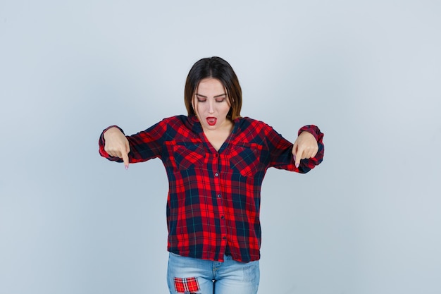 Mujer joven en camisa a cuadros, jeans apuntando hacia abajo con los dedos índices, manteniendo la boca abierta y mirando sorprendido, vista frontal.
