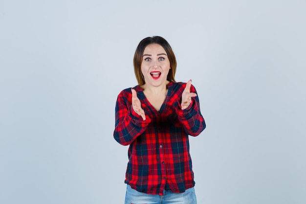 Mujer joven en camisa a cuadros, jeans estirando las manos hacia y mirando sorprendido, vista frontal.