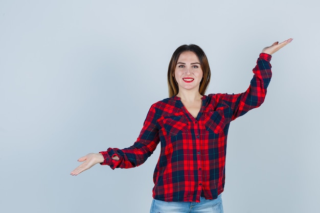 Mujer joven en camisa a cuadros mostrando gesto de escalas y mirando alegre, vista frontal.