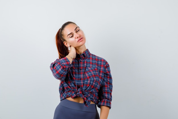 Mujer joven en camisa a cuadros, pantalones con la mano en el cuello y mirando exhausto, vista frontal.