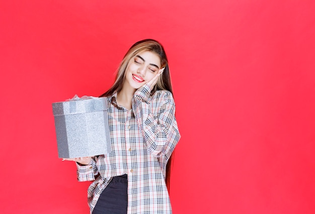 Mujer joven en camisa a cuadros sosteniendo una caja de regalo plateada y parece relajada y somnolienta
