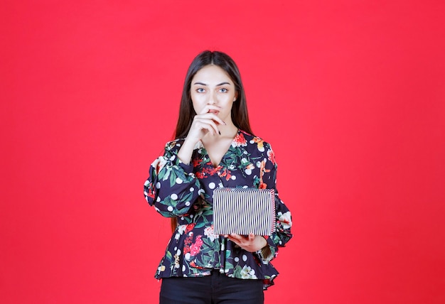 Mujer joven en camisa floral sosteniendo una caja de regalo plateada y parece pensativa