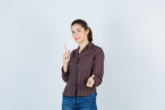 Mujer joven en camisa a rayas, jeans apuntando hacia arriba con el dedo índice, mostrando el pulgar hacia arriba y mirando alegre, vista frontal.