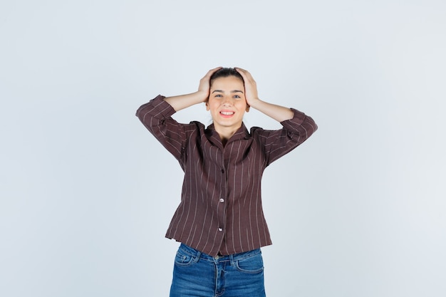 Mujer joven en camisa a rayas, jeans con las manos en la cabeza, sonriendo y mirando alegre, vista frontal.