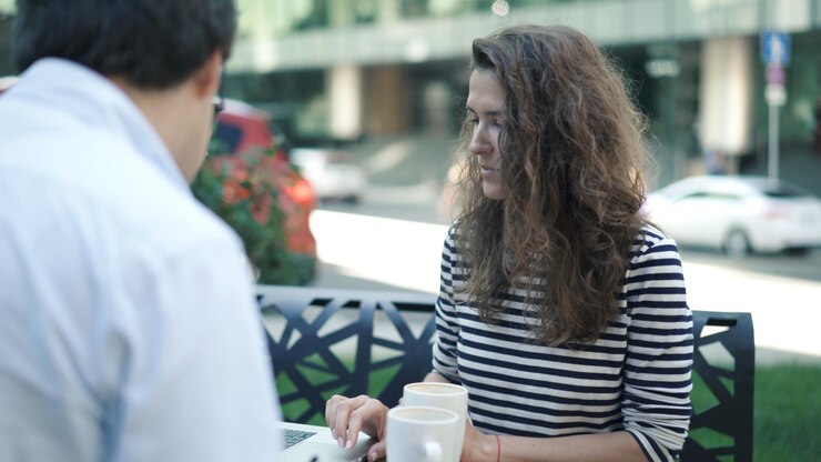 Mujer joven con camisa a rayas y su apuesto colega con camisa blanca trabajando juntos en el parque de verano usando una computadora portátil y un portapapeles El hombre está tomando café