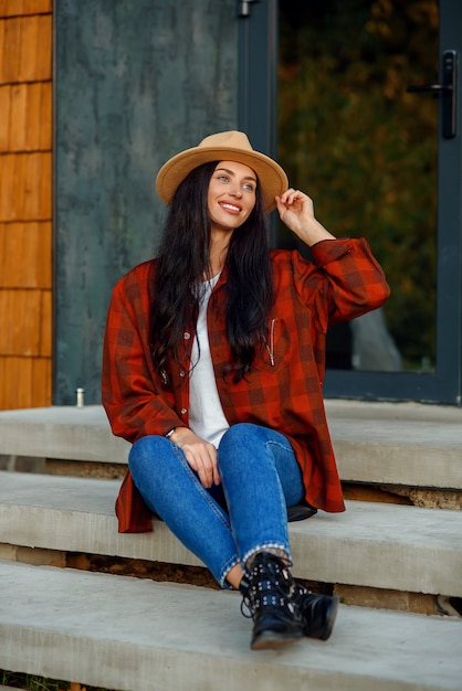 La mujer joven en una camisa roja de moda sombrero marrón y pantalones de mezclilla se sienta en una escalera, cerca de su propia casa.