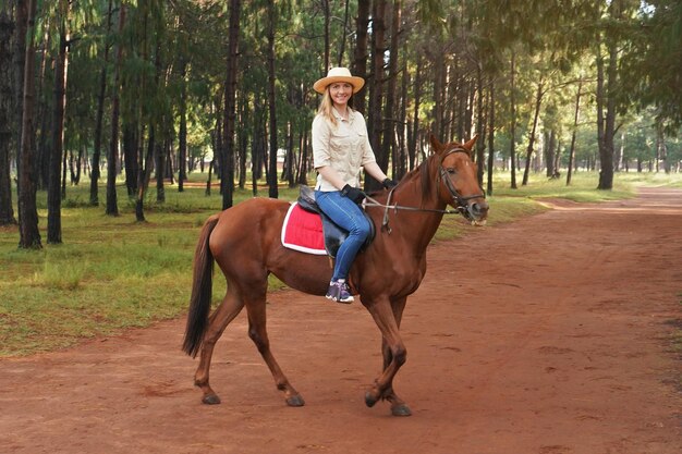 Mujer joven con camisa y sombrero de paja, montando caballo marrón, fondo borroso con árboles en el parque