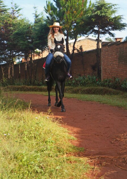 Mujer joven con camisa y sombrero de paja, montando un caballo negro en el parque sobre un camino de tierra, una pared de ladrillo cerca de ella, en una mañana soleada