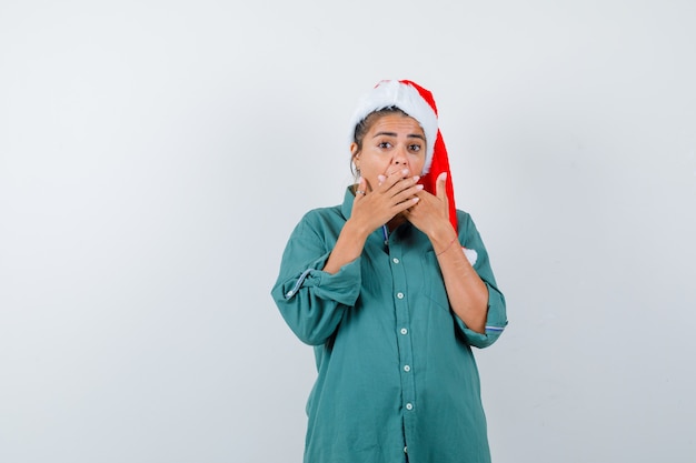 Mujer joven en camisa, sombrero de Santa Claus cubriendo la boca con las manos y mirando horrorizado, vista frontal.