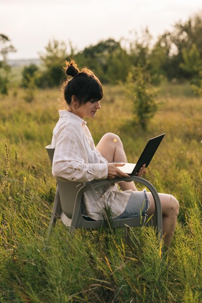 Una mujer joven con una camisa trabaja en una computadora portátil mientras está sentada en una silla en medio de la naturaleza