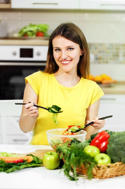 Mujer joven en camiseta amarilla revolviendo ensalada de verduras en un recipiente transparente tiro interior en cocina blanca moderna