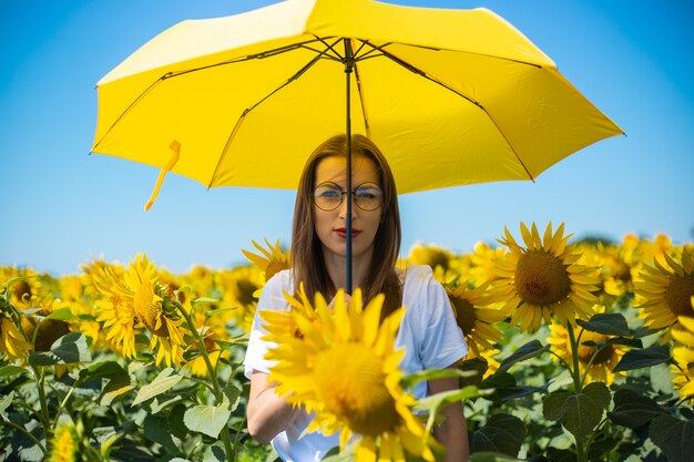 Mujer joven en camiseta blanca y gafas bajo un paraguas amarillo en un campo de girasoles.