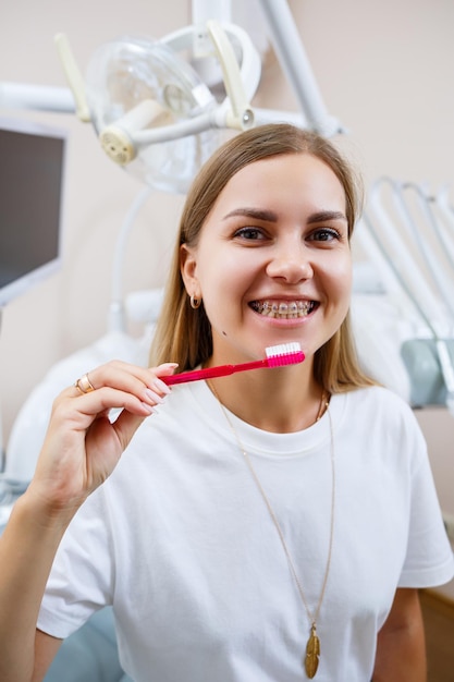 Una mujer joven con una camiseta blanca se sienta en un consultorio dental, sonríe y sostiene un cepillo de dientes en sus manos. Chica con aparatos ortopédicos muestra cuidado bucal. Odontología, tratamiento dental