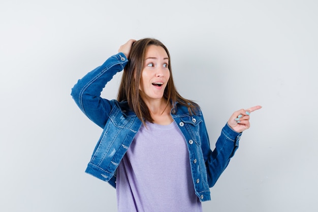 Mujer joven en camiseta, chaqueta con la mano en la cabeza, apuntando hacia la derecha y mirando sorprendido, vista frontal.