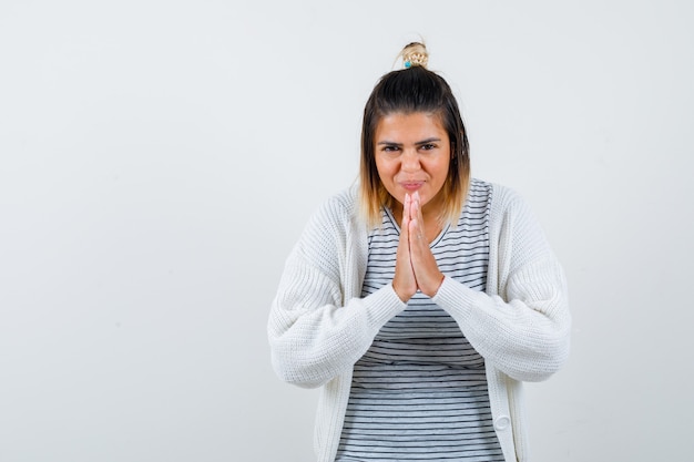 Mujer joven en camiseta, chaqueta de punto mostrando gesto de namaste y mirando agradecido, vista frontal.