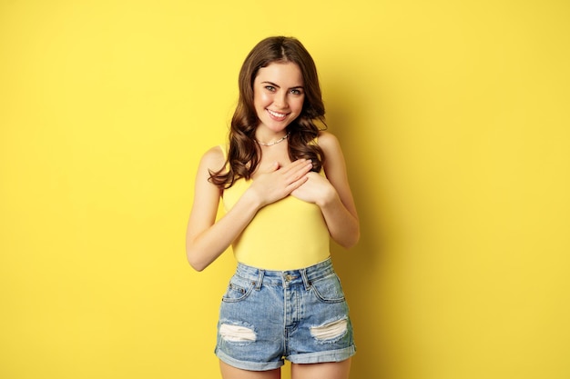 Mujer joven en camiseta sin mangas, tomándose de la mano en el corazón con cuidado y ternura, sonriendo a la cámara, luciendo agradecida, agradecida, de pie sobre un fondo amarillo.