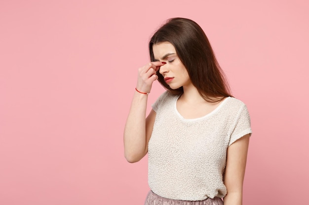 Mujer joven cansada con ropa ligera informal posando aislada en un fondo rosa, retrato de estudio. Gente emociones sinceras concepto de estilo de vida. Simulacros de espacio de copia. Manteniendo los ojos cerrados, poniendo la mano en la nariz.