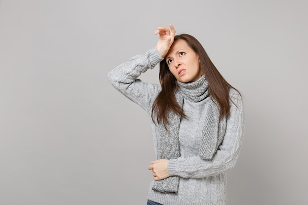 Mujer joven cansada en suéter gris, bufanda poniendo la mano en la cabeza, mirando hacia arriba aislado sobre fondo de pared gris. Gente de estilo de vida de moda saludable emociones sinceras concepto de estación fría. Simulacros de espacio de copia.