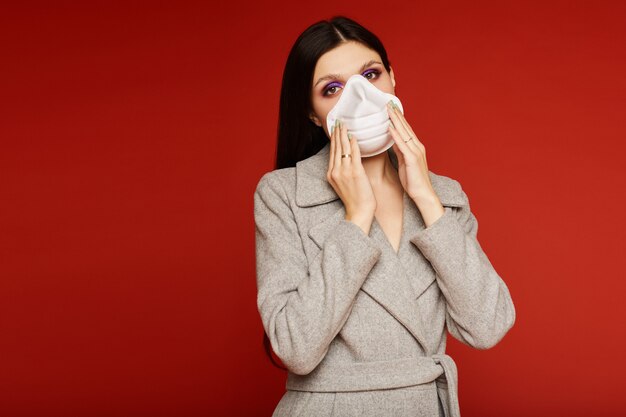 Mujer joven en capa y respirador que mira en la cámara, aislada. Concepto de protección contra la epidemia de gripe, alergia al polvo y contaminación del aire.