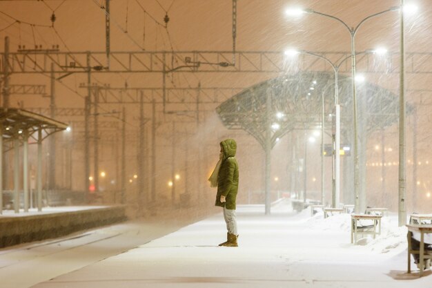 Mujer joven en el capó en la plataforma ferroviaria vacía en Blizzard esperando un tren.