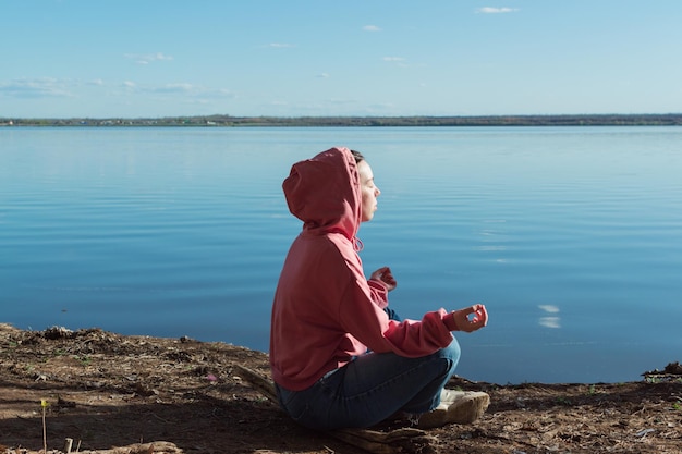 Una mujer joven con capucha se sienta en la orilla de un lago con los ojos cerrados y medita.