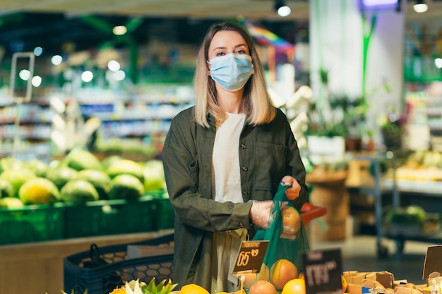 Mujer joven en la cara máscara protectora médica elige y recoge en bolsa ecológica verduras o frutas en el supermercado Mujer de pie en un mercado de la tienda de comestibles cerca del mostrador compra en un paquete reutilizable