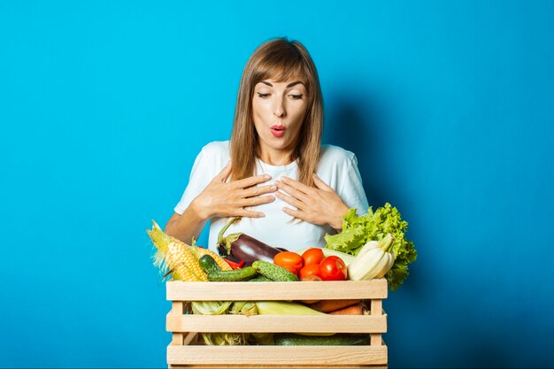 La mujer joven con una cara sorprendida sostiene una caja con las verduras frescas en azul. Buen concepto de cosecha, producto natural.