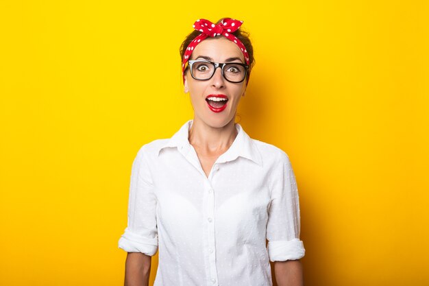 Mujer joven con cara de sorpresa, con gafas y una diadema en la cabeza sobre una pared amarilla.