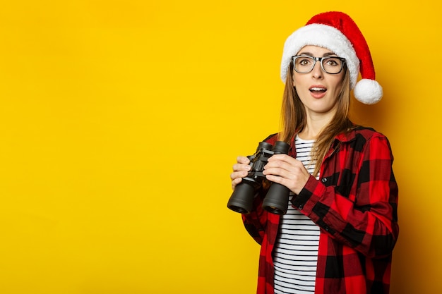 Mujer joven con una cara de sorpresa y con un gorro de Papá Noel y una camisa roja en una jaula tiene binoculares