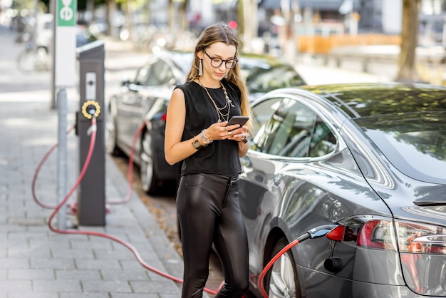 Mujer joven cargando un coche eléctrico de pie con un teléfono inteligente al aire libre en la calle en la ciudad de Rotterdam