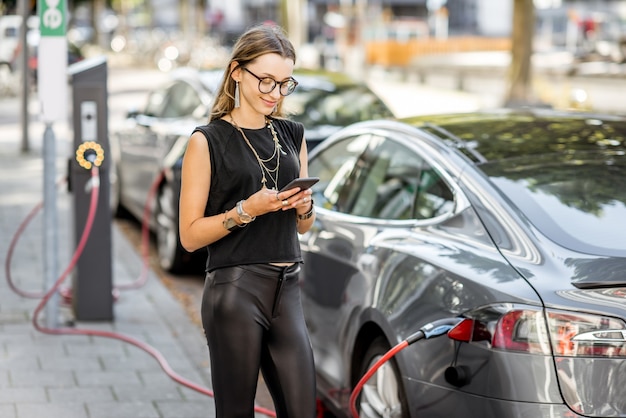 Mujer joven cargando un coche eléctrico de pie con un teléfono inteligente al aire libre en la calle en la ciudad de Rotterdam