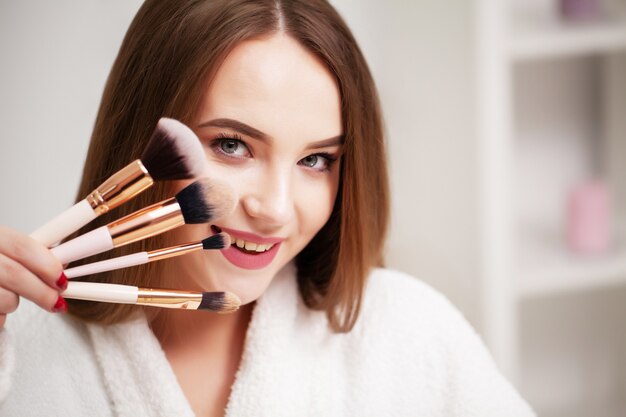 Mujer joven en casa aplica maquillaje en la cara en el baño frente a un espejo.